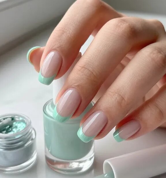 Ultra-realistic close-up beauty photo of Short Green nails featuring a “Chrome Mint Micro-French” manicure: short, natural-length nails with a sheer glossy nude base and ultra-thin mint green chrome tips that have a mirrored sheen; shot on a well-groomed hand holding a minimal glass bottle on a marble vanity. Include visible nail tools: a fine detail brush with mint gel, a small jar of chrome powder, tip guides, a sheer nude polish bottle, and a glossy top coat. Soft daylight from a window, clean modern aesthetic, neutral background, shallow depth of field to showcase the precision of the skinny mint chrome line and high-gloss finish. Skin tone natural, cuticles hydrated, no chips, realistic texture and reflections, beauty editorial quality.