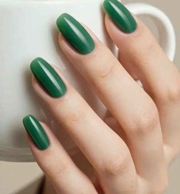 Ultra-realistic close-up beauty photo of a hand with perfectly manicured almond-shaped nails painted in deep Jade Green, captured under soft studio lighting on a neutral beige background. The nails feature a mirror-like, high-gloss finish that mimics polished jade stone, with subtle tonal depth and natural highlights. Clean, precise cuticle work emphasizes the rich Jade Green color and a refined, expensive look. The almond shape is balanced and not too pointy, with softly rounded tips that elongate the fingers. Include a delicate ceramic espresso cup held between fingers to suggest a chic coffee date, and a blurred hint of a minimalist blazer cuff for a boardroom-ready vibe. High resolution, macro detail on nail surface and cuticles, natural skin texture, no harsh retouching, realistic color rendering showcasing true Jade Green.